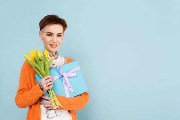 Mujer alegre con tatuajes con tulipanes amarillos y caja de regalo envuelta aislada en azul - foto de stock