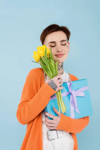 Femme heureuse avec des tatouages tenant des tulipes jaunes et boîte cadeau enveloppé isolé sur bleu — Photo de stock