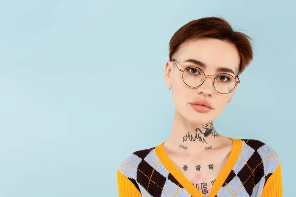 Молода татуйована жінка в окулярах дивиться на камеру ізольовано на синьому — стокове фото