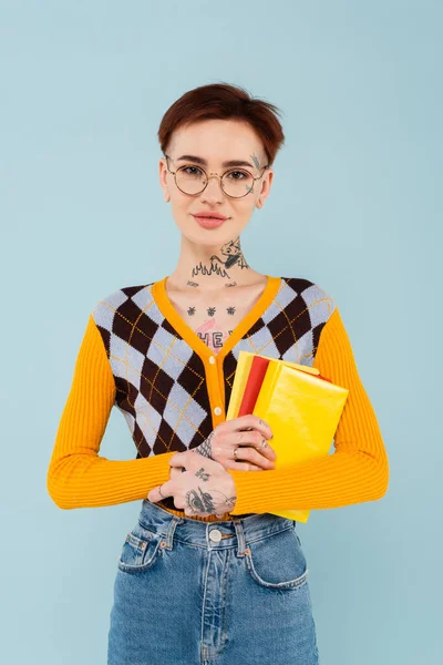 Estudante tatuado alegre em óculos segurando livros isolados em azul — Fotografia de Stock