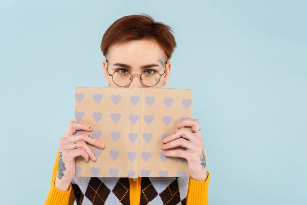 Mujer joven tatuada en gafas que cubren la cara con libro en la cubierta del patrón del corazón aislado en azul - foto de stock