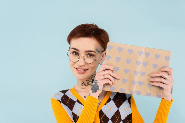 Happy tattooed woman in glasses holding book in heart pattern cover isolated on blue — Stock Photo