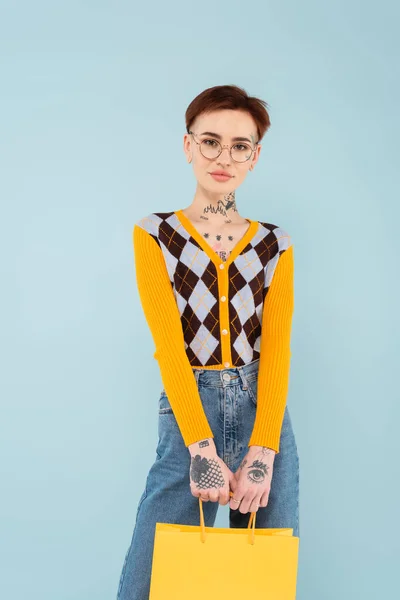 Jeune femme tatouée dans des lunettes et cardigan tenant sac à provisions isolé sur bleu — Photo de stock