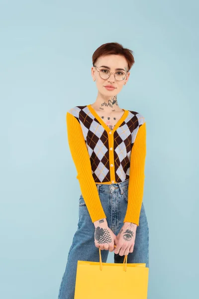 Mujer joven tatuada en gafas y cárdigan sosteniendo bolsa aislada en azul - foto de stock