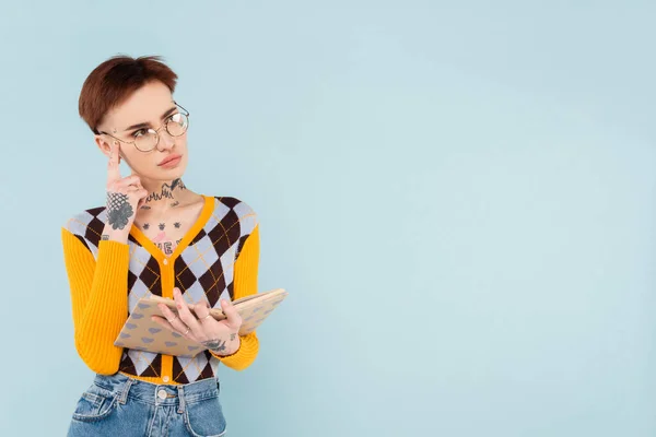 Tätowierte Frau in Brille mit Buch im Herzmuster-Einband und Denken isoliert auf blau — Stockfoto