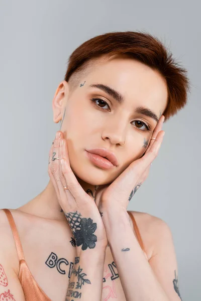 Young tattooed woman looking at camera and touching face isolated on grey — Stock Photo