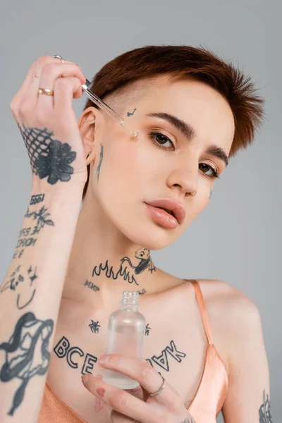 Giovane donna tatuata che tiene bottiglia e pipetta mentre applica siero sul viso isolato su grigio — Stock Photo