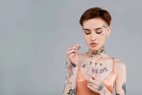 Mujer joven con tatuajes sosteniendo botella con suero y pipeta aislados en gris - foto de stock