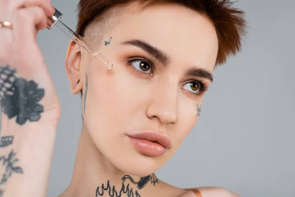 Giovane donna tatuata che applica siero con pipetta isolata su grigio — Stock Photo