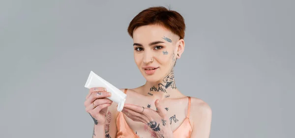 Mujer joven tatuada sosteniendo tubo y aplicando loción corporal mientras sonríe aislada en gris, pancarta - foto de stock