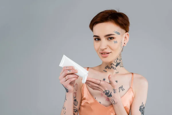 Tatuato giovane donna in possesso di tubo e l'applicazione di lozione per il corpo, mentre sorridente isolato su grigio — Foto stock