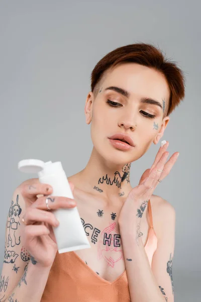 Young woman with tattoos holding white tube and applying face cream isolated on grey — Stock Photo