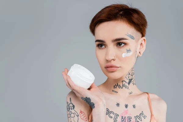 Giovane donna con tatuaggi e capelli rossi in possesso di contenitore bianco con prodotto cosmetico isolato su grigio — Foto stock