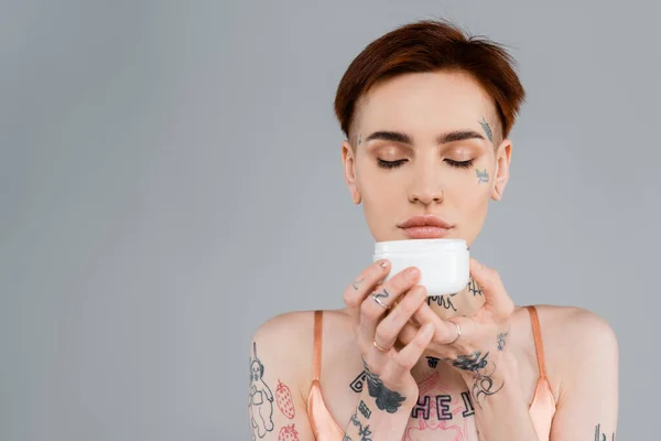 Young woman with tattoos and red hair holding white container while smelling cosmetic product isolated on grey — Stock Photo