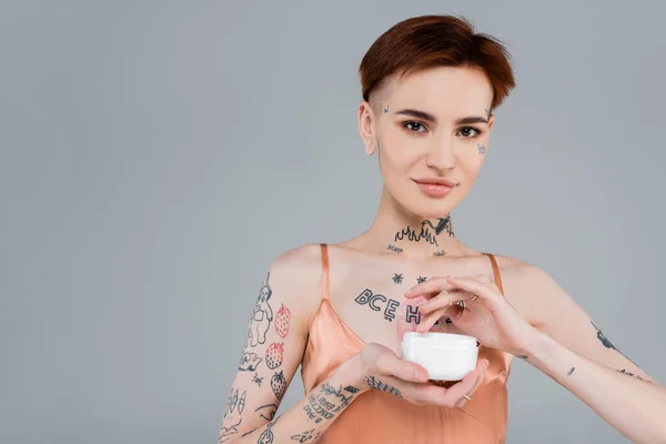 Mujer feliz con tatuajes y pelo rojo sosteniendo contenedor blanco con producto cosmético aislado en gris - foto de stock