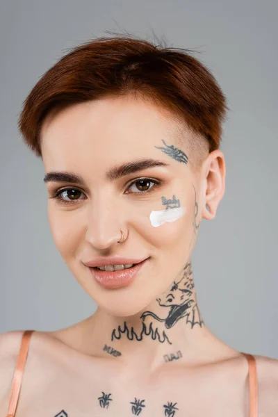 Femme tatouée et heureuse avec crème cosmétique sur le visage souriant isolé sur gris — Photo de stock