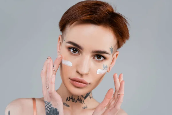 Jeune femme tatouée appliquant crème hydratante sur le visage isolé sur gris — Photo de stock