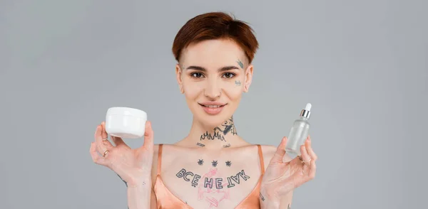 Mujer joven y tatuada sosteniendo productos cosméticos mientras sonríe aislada en gris, pancarta - foto de stock