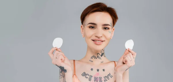 Mujer feliz y tatuada sosteniendo almohadillas de algodón aisladas en gris, pancarta - foto de stock