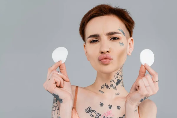 Joven mujer tatuada haciendo pucheros labios y sosteniendo almohadillas de algodón aisladas en gris - foto de stock