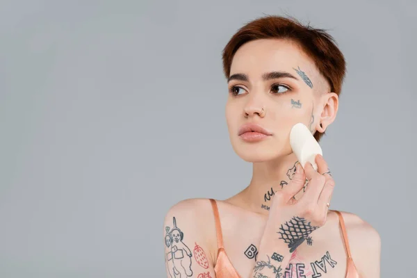 Tattooed woman applying makeup foundation on face with sponge isolated on grey — Stock Photo