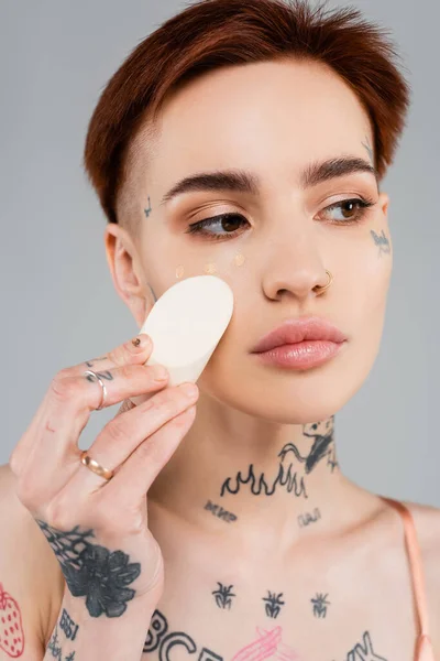 Tattooed woman applying makeup foundation with sponge isolated on grey — Stock Photo