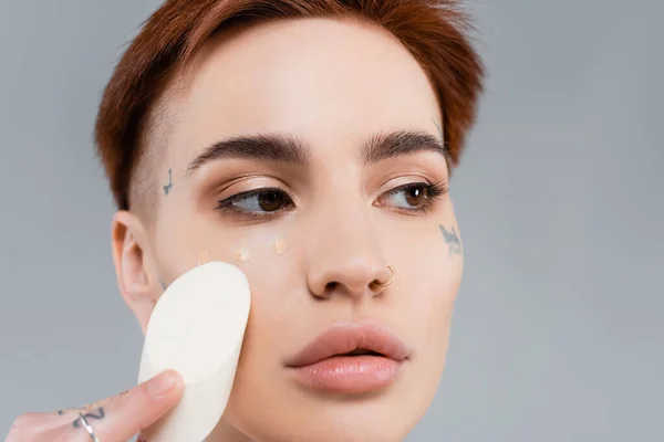 Close up de mulher tatuada aplicando base de maquiagem no rosto com esponja isolada em cinza — Fotografia de Stock
