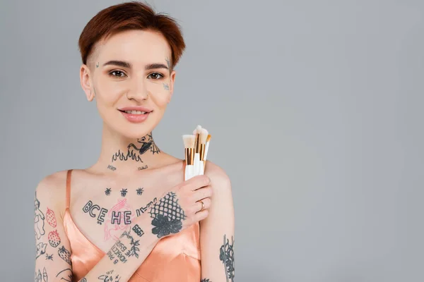 Счастливая и молодая татуированная женщина с косметическими кисточками, изолированными на сером — стоковое фото