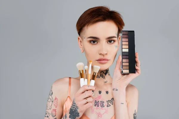 Young tattooed woman holding eye shadow palette and cosmetic brushes isolated on grey — Stock Photo