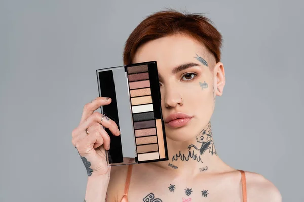 Mujer joven y tatuada sosteniendo la paleta de sombras oculares aislada en gris - foto de stock