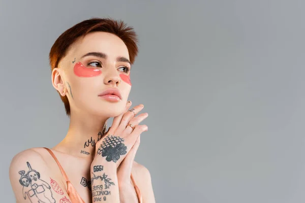 Jovem mulher com tatuagens em manchas olho hidratante olhando para longe isolado em cinza — Fotografia de Stock