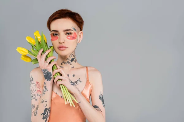 Молодая и татуированная женщина в розовых повязках на глазу держит тюльпаны изолированные на сером — стоковое фото