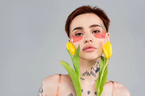 Tattooed woman in pink eye patches holding yellow tulips isolated on grey — Stock Photo