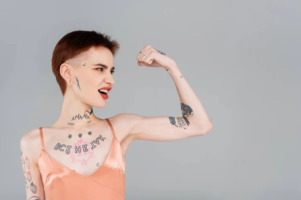 Jeune femme tatouée avec des lèvres rouges montrant le muscle isolé sur gris — Photo de stock