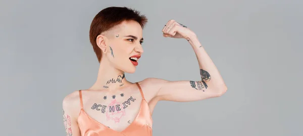Tattooed young woman with red lips showing muscle isolated on grey, banner — Stock Photo