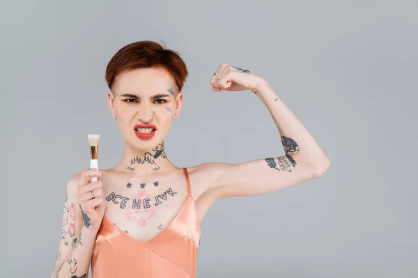 Tatuato giovane donna con labbra rosse mostrando muscoli e tenendo pennello cosmetico isolato su grigio — Foto stock