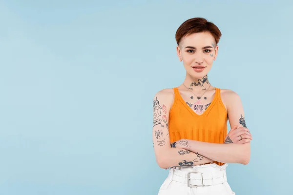 Mujer complacida y tatuada en camiseta de punto naranja con brazos cruzados aislados en azul - foto de stock