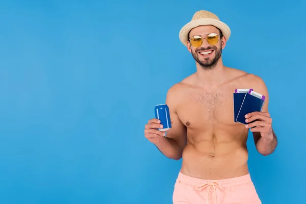 Fröhlicher, hemdloser Mann mit Sonnenbrille, Getränkedosen und Reisepässe isoliert auf blauem Grund — Stockfoto