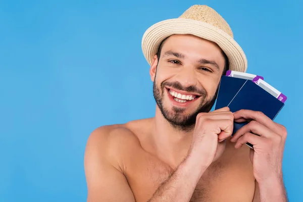 Hombre sin camisa alegre con sombrero de sol sosteniendo pasaportes con billetes de avión aislados en azul - foto de stock
