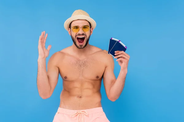 Hombre sorprendido en gafas de sol con pasaportes y billetes de avión aislados en azul - foto de stock