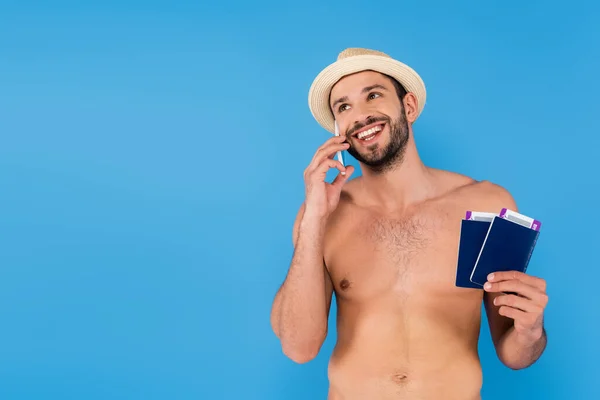 Alegre hombre sin camisa hablando en el teléfono inteligente y la celebración de pasaportes con billetes de avión aislados en azul - foto de stock