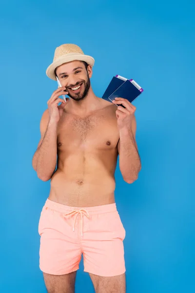 Alegre hombre sin camisa hablando en el teléfono inteligente y la celebración de pasaportes aislados en azul - foto de stock