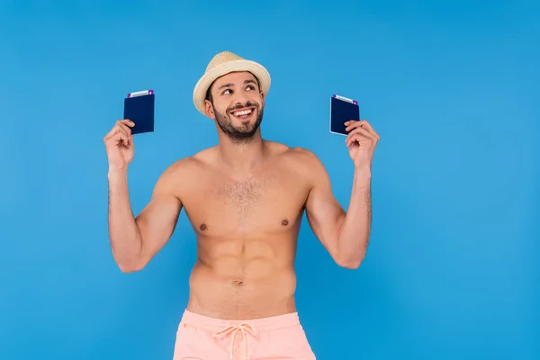 Positivo uomo senza camicia con cappello da sole in possesso di passaporti e biglietti aerei isolati su blu — Foto stock