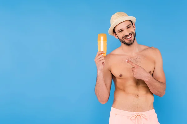 Hemdloser Mann mit Sonnenhut zeigt auf Sonnencreme isoliert auf blau — Stockfoto