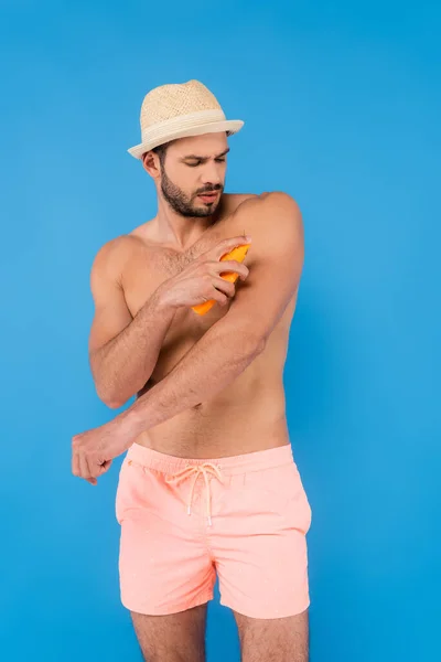 Homem sem camisa em chapéu de sol aplicando protetor solar isolado em azul — Fotografia de Stock