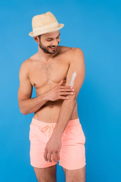 Homem sem camisa sorridente em chapéu de palha aplicando protetor solar isolado em azul — Fotografia de Stock