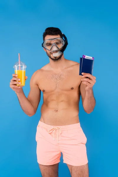 Shirtless man in swimming goggles holding passports and orange juice isolated on blue — Stock Photo