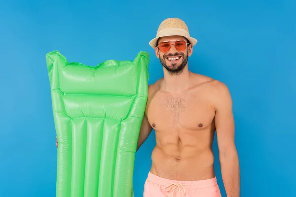 Homem sem camisa em chapéu de sol e óculos de sol segurando colchão inflável isolado em azul — Fotografia de Stock
