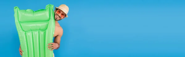 Hombre alegre en gafas de sol y sombrero de sol que sostiene el colchón inflable aislado en azul - foto de stock