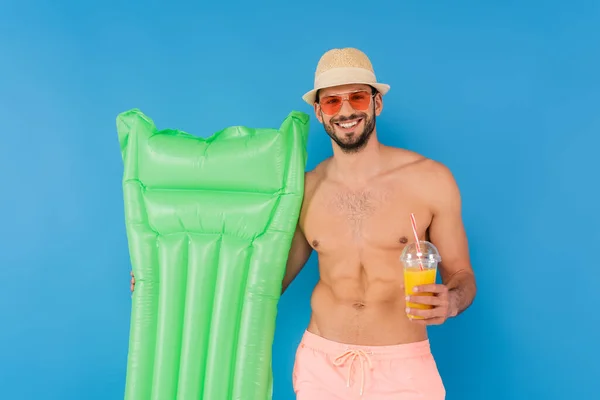 Smiling man in sunglasses holding orange juice and inflatable mattress isolated on blue — Stock Photo